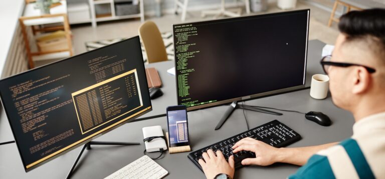 Mężczyzna piszący na klawiaturze przy dwóch monitorach wyświetlających kod i smartfonie na biurku – shadow ai a szbi